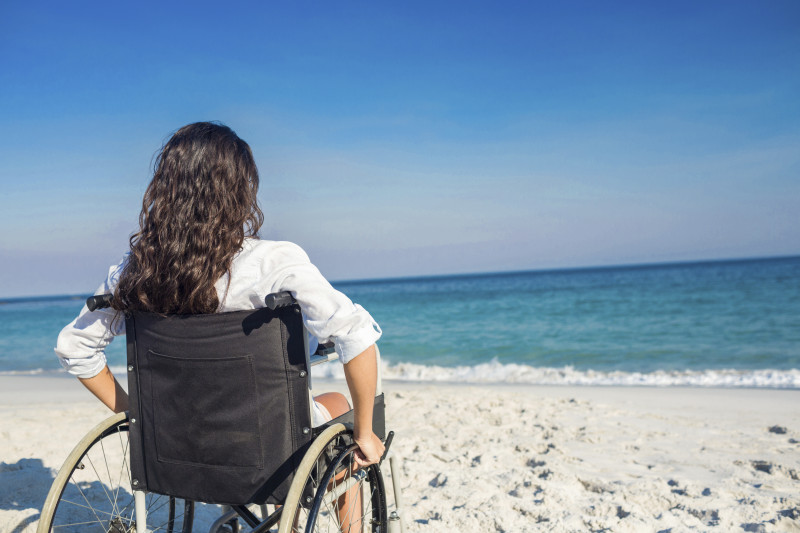 A young woman seated in a wheelchair on the beach, gazing at the ocean horizon.