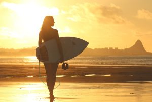 Silhouette of a woman walking on the beach carrying a surfboard at sunset.